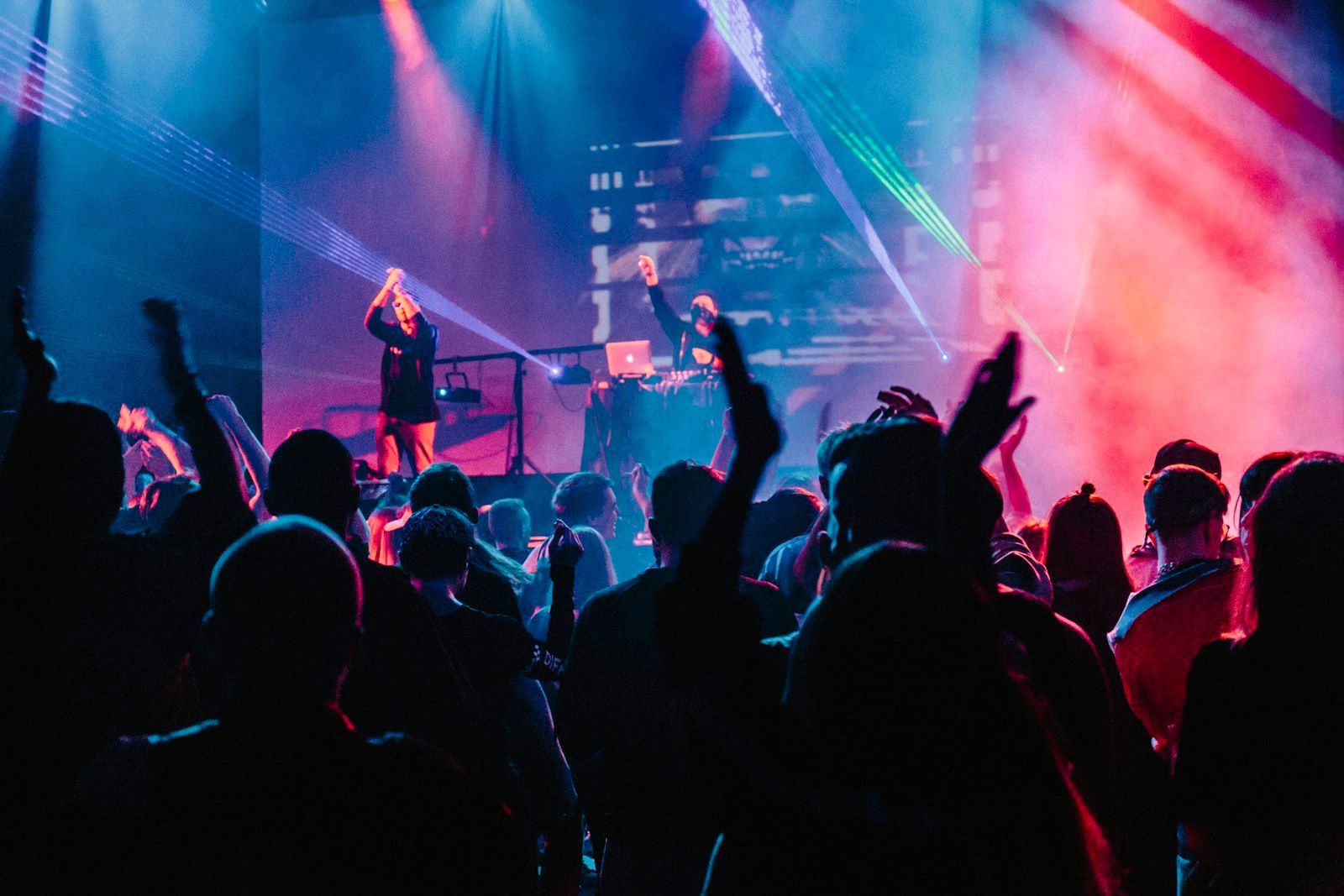 Silhouetted crowd with raised hands facing a DJ on a stage lit by pink and blue lasers