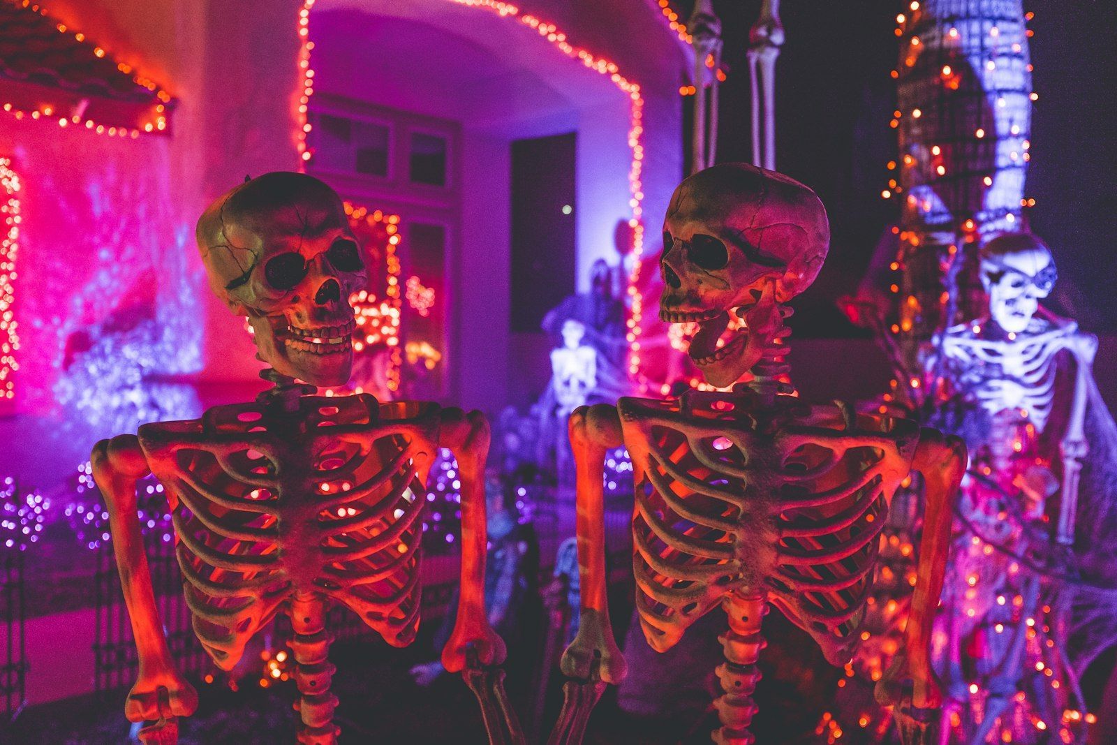 Two skeleton decorations in front of a house lit with purple and orange Halloween lights at night