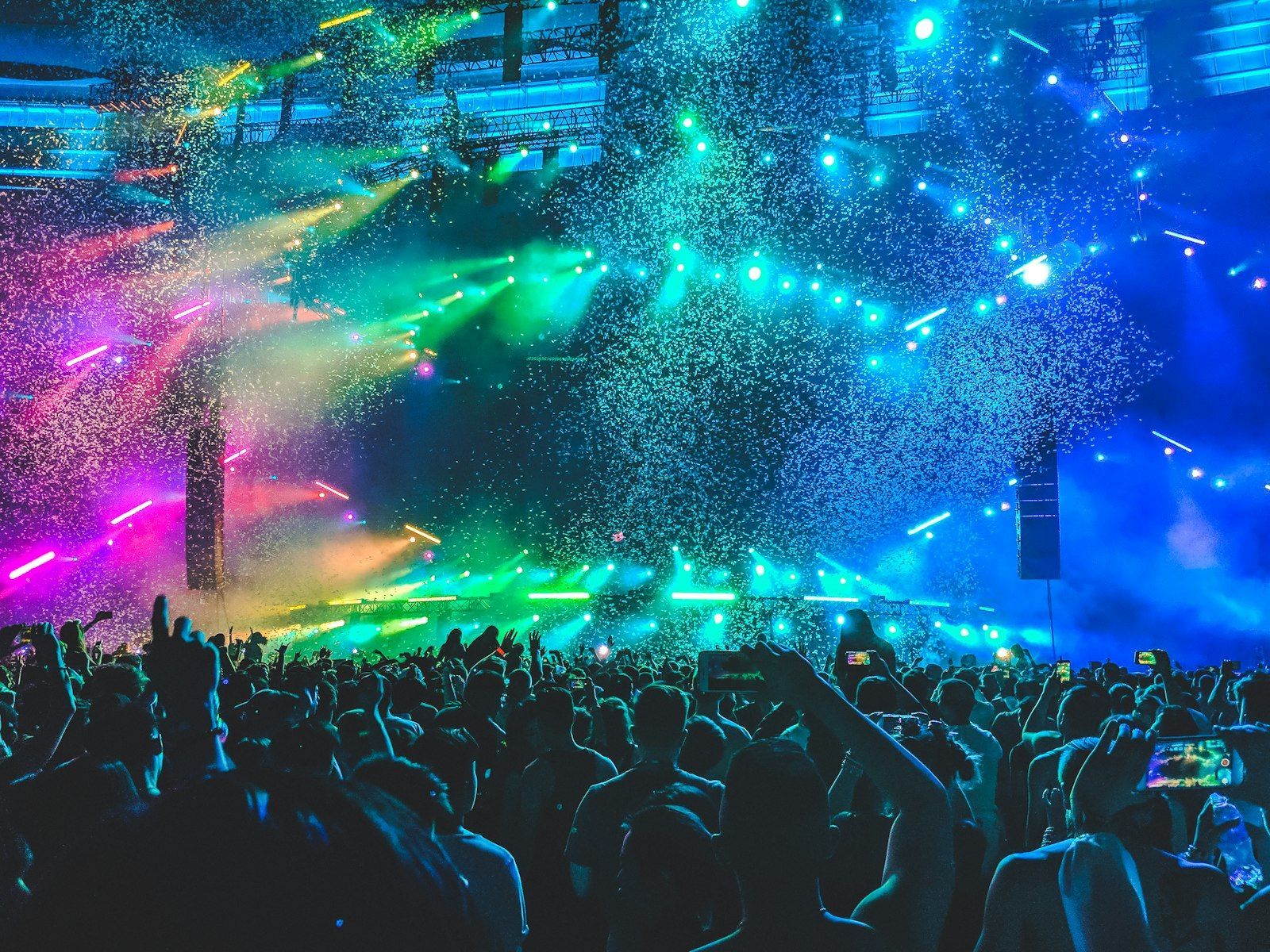 Festival crowd with raised hands and phones under confetti and green, blue and pink stage lights