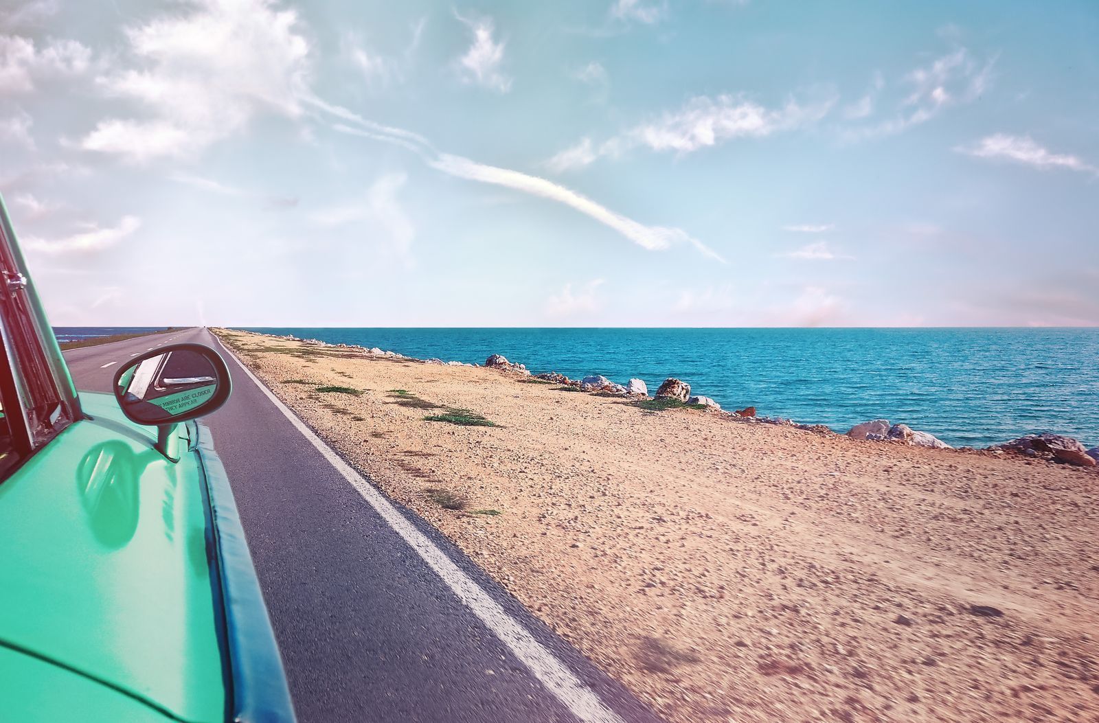 Vintage green car driving along a coastal road beside a beach and turquoise sea