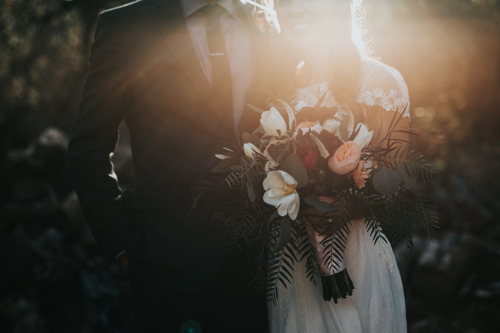 A groom in a dark suit holding a bridal bouquet of white and peach flowers in warm sunlight